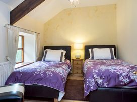 A bedroom with two single beds with floral purple bedspreads a wooden nightstand with a lamp and a window with white curtains at Mill Cottage in Aylesbeare