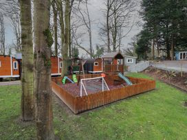 A fenced playground with a slide swings and a small climbing structure surrounded by trees and cabins at Hexagon Hut - Dec Update in Dungiven Londonderry