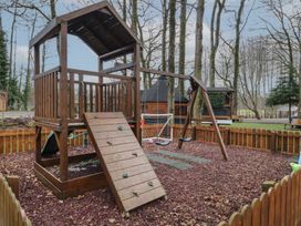 A wooden playground structure with a climbing wall swing set and slide in a fenced outdoor area surrounded by trees at Hawthorn in Dungiven Londonderry