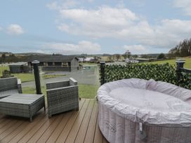 A deck with a covered hot tub and wicker chairs overlooking a rural landscape at Debonair Lodge in Three Lochs Holiday Park near Glenluce