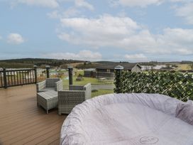 An outdoor deck with wicker chairs a round covered hot tub and a view of a residential area with houses and trees at Debonair Lodge in Three Lochs Holiday Park near Glenluce
