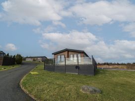 A lodge with a fenced porch on a grassy hill near a paved road at Debonair Lodge in Three Lochs Holiday Park near Glenluce