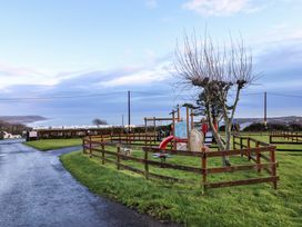 A fenced playground with slides and climbing equipment on grass near a paved path with sea and hills in the background at Chalet 27 in Port Eynon
