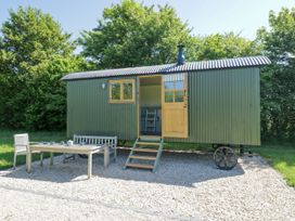 A green corrugated metal shepherd hut with open door and window on a gravel patio with wooden table chairs and bench surrounded by trees at Hazel South Leigh near Witney