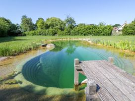An outdoor natural swimming pond with a wooden dock in a grassy area with trees and a house in the background at Hazel South Leigh near Witney