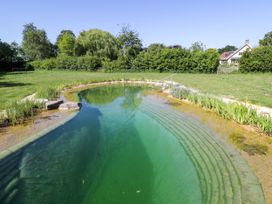 An outdoor natural swimming pool with stone steps and surrounding plants in a grassy area with trees and a house in the background at Spindle South Leigh near Witney