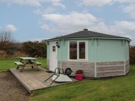 A small round cottage with a white door and window next to a wooden picnic table on a deck with a wheelbarrow and a pink flower pot outside Blackpool Mill near Hartland