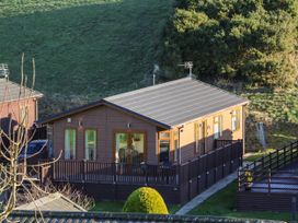 A wooden cabin with a fenced porch and green hillside in the background at The Sanctuary in Badgers Retreat Holiday Park near Catterick Garrison North Yorkshire