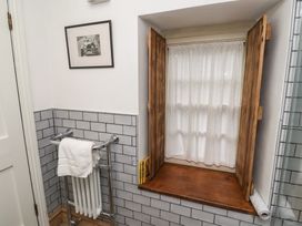 A bathroom window with wooden shutters and white curtains next to a towel rack with a white towel and a black and white picture on the tiled wall at The Old Coach House in Eglingham near Alnwick