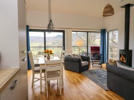 A dining table with chairs and flowers next to sofas and a wood stove in a living room with large windows at Dundarach in Aberfeldy