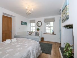 A bedroom with a bed covered with white and beige bedding two folded towels on the bed wooden door two windows with roman blinds white cabinet round mirror armchair gray rug and a fireplace with floral arrangement at Pippin Cottage in Beaumaris