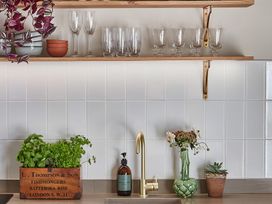 A kitchen counter with a wooden planter of herbs soap dispenser gold faucet flower vase and potted succulent under wooden shelves with glasses at The Stables Galteemore Farm in Avebury near Marlborough