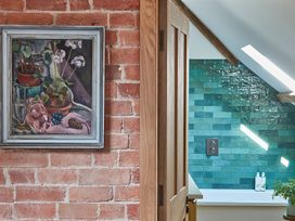 A bathroom with green tiled wall and bottles by a bathtub viewed through a wooden door with a brick wall and a framed painting nearby at The Stables in Galteemore Farm in Avebury near Marlborough