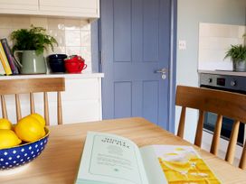 A kitchen table with a bowl of lemons and an open recipe book near wooden chairs a blue door a countertop with stacked bowls cups and potted plants and a built-in oven at The Stables in Galteemore Farm in Avebury near Marlborough