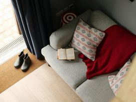 A gray sofa with patterned cushions and a red blanket an open book on the sofa green boots near a curtain at The Stables Galteemore Farm in Avebury near Marlborough