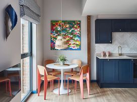 A dining area with a white round table and four wooden chairs with pink legs next to a kitchen with blue cabinets and a wooden floor at The Stables in Galteemore Farm in Avebury near Marlborough