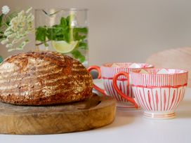 A loaf of bread on a wooden board with two patterned teacups and a glass jug with lime and mint leaves