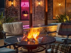 An outdoor seating area with cushioned chairs around a fire pit table with cups and blankets near a brick wall with a neon sign at The Stables Galteemore Farm in Avebury near Marlborough