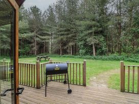 A wooden deck with a black grill and a picnic table on grass near trees at Rowan Lodge in Rosliston