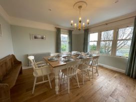 A dining room with a wooden table set with plates and bowls surrounded by white chairs and a wooden bench near windows with curtains at Bryn Awelon in Harlech