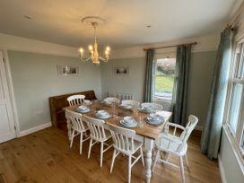 A dining room with a wooden table set for eight people with white chairs and windows with curtains at Bryn Awelon in Harlech