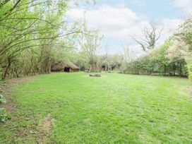 A grassy yard with small thatched huts and a fire pit surrounded by trees at Brandon House Farm Yurt in Brandon Parva near Mattishall