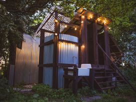An outdoor structure with corrugated metal walls wooden frame string lights and a sink surrounded by trees and plants at Brandon House Farm Yurt Brandon Parva near Mattishall