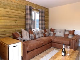 A corner sofa with cushions next to a window with curtains and a wooden table with a bottle and glasses at Robins Nest Lodge in Snettisham