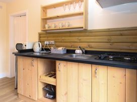 A kitchen counter with wooden cabinets a sink a stovetop a kettle a microwave and shelves with mugs and glasses at Robins Nest Lodge in Snettisham