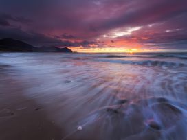 A beach with waves and a sunset sky with clouds and mountains in the background