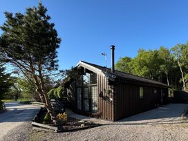 A single-story dark wooden house with a sloped roof and a tall window next to a gravel driveway at Fig Tree Lodge in Hutton Roof near Penrith