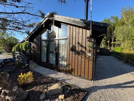 A brown wooden cabin with large windows and a gravel pathway surrounded by trees and shrubs at Fig Tree Lodge in Hutton Roof near Penrith