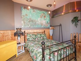 A bedroom with a black metal bed frame and tropical patterned bedding a yellow chest a small white side table with a lamp an ironing board on a black rack and a wooden chair shaped like a hand at Mermaids Rest in Newquay