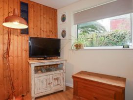 A corner of a room with a TV on a wooden cabinet a floor lamp a wooden chest a window with a plant on the sill and framed pictures on the wall at The Aqua Shack in Newquay