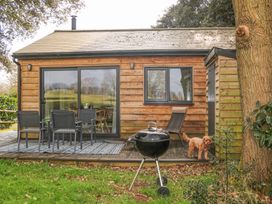 A wooden cottage exterior with a deck table and chairs a barbecue grill and a dog on the grass at Holm Oaks Cottage in Busbridge near Godalming