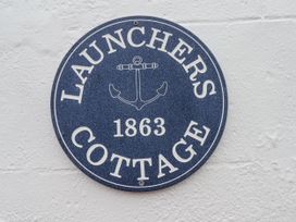 A round blue sign with an anchor icon and text that reads Launchers Cottage 1863 mounted on a white wall