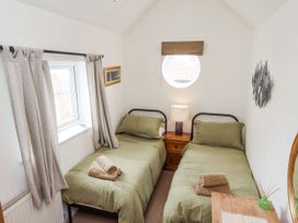 A small bedroom with two single beds with green bedding and folded towels a wooden nightstand between the beds with a lamp a round window with a brown blind curtains on a rectangular window on the left and wall decorations at Launchers Cottage in Scarborough