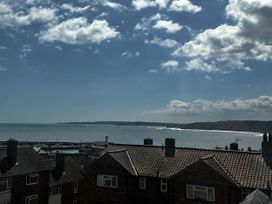 A coastal view showing rooftops of houses in front of a harbor with a lighthouse and boats plus a body of water and distant cliffs under a partly cloudy sky at Launcher's Cottage in Scarborough