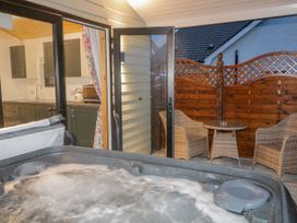 An outdoor patio with a hot tub and two wicker chairs and a small round table near a wooden fence with open doors leading to a kitchen at Benone Cabin in Magilligan near Castlerock