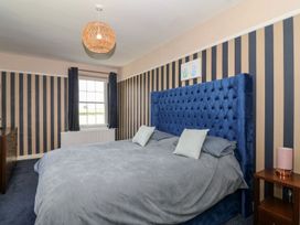 A bedroom with a large blue bed, striped wallpaper, a window with curtains, a chest of drawers and a bedside table with a lamp at Buckland House and Annex Lower Durston near Taunton