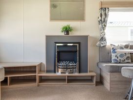 A living room with a fireplace a wooden shelf a plant a window with curtains and a gray sofa with patterned cushions at Ripples & Reeds - Billing Aquadrome