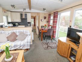 An open living area with a plaid sofa, a wooden coffee table with flowers, a dining table set with plates and glasses, a kitchen with cabinets and appliances, and a television on a wooden stand at The Retreat Near Shorwell