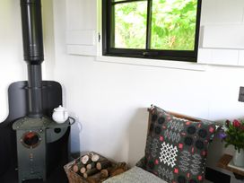 A wood burning stove with a white teapot on top next to a basket of firewood a patterned cushion on a chair and a window showing green foliage at Bugail Hut in Llantwit Major