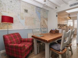 A dining area with a wooden table and distressed white chairs next to a red plaid armchair and a floor lamp by a wall with maps at Lychgate View Cottage in Aldbrough