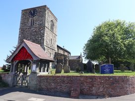 A stone church with a clock tower and a wooden lychgate covered in red flowers at Lychgate View Cottage in Aldbrough