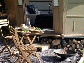 An outdoor dining area with a wooden table and chairs a small fire pit and logs stacked near a cabin with an open door showing a bed inside at Caban Llwynon Tresillian Wood near Llantwit Major