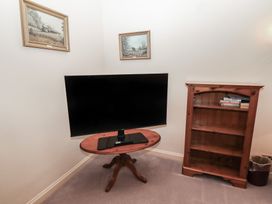 A corner of a room with a flat screen tv on a wooden oval table a wooden bookshelf with some books and a trash bin