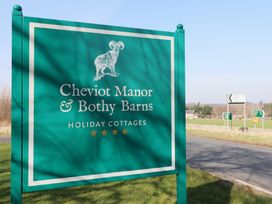 A green sign for Cheviot Manor and Bothy Barns holiday cottages by a roadside with distance signs in the background at Bizzie Lizzie in Akeld near Wooler