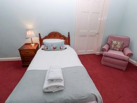 A single bed with a floral pillow and folded towels on red carpet next to a wooden nightstand with a lamp and a pink armchair with a floral cushion at Cheviot Manor Akeld near Wooler