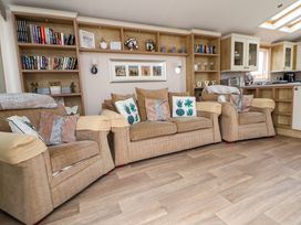 A living room with three beige armchairs, cushions with floral patterns, built-in bookshelves with books and decorative items, and a kitchen area in the background at Hazel Park Lodge HP4 in Rhyl